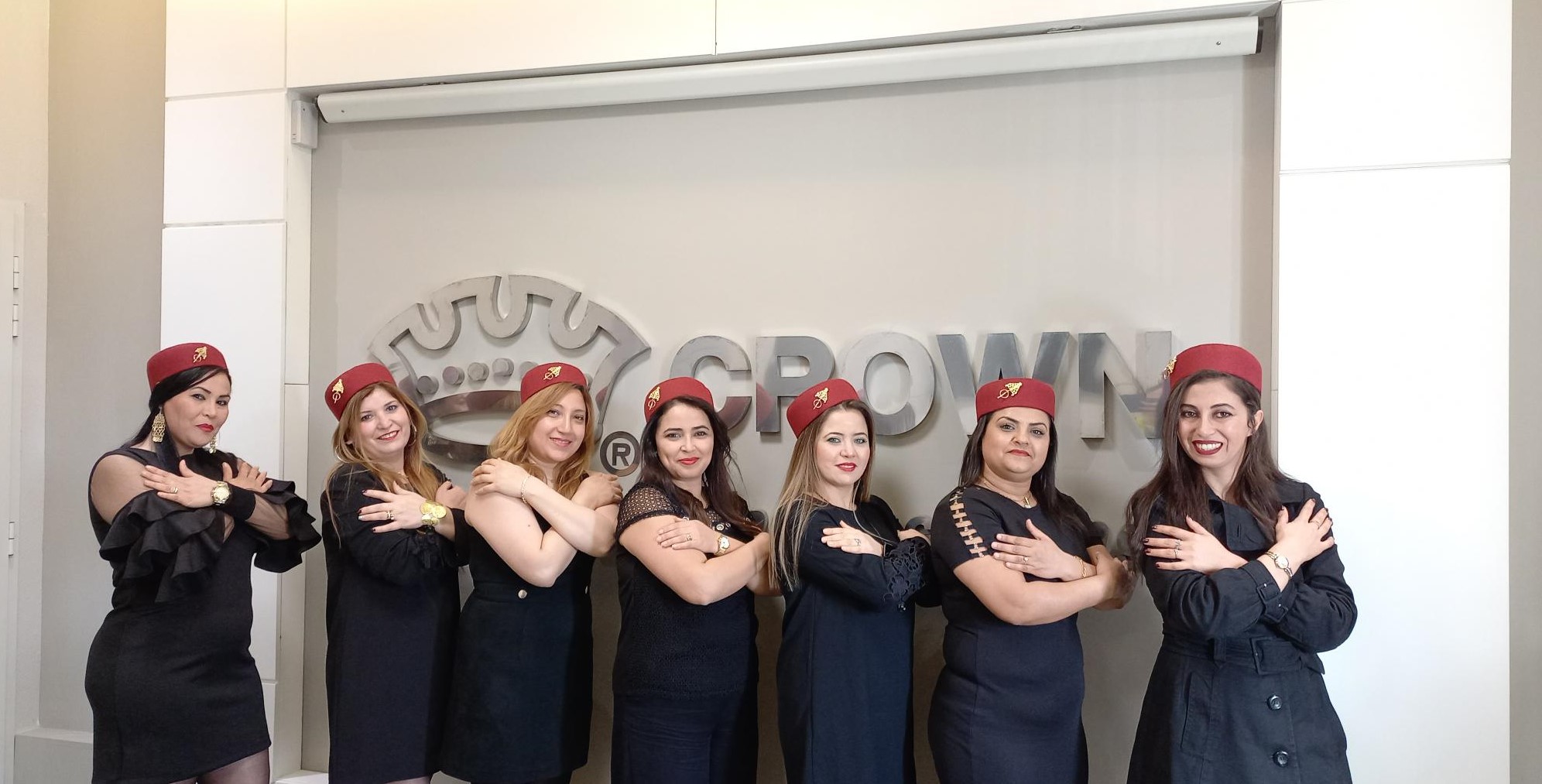A group of women in navy dresses and red hats stand in front of the Crown Cork logo. They have their arms crossed in the symbol for 'equity'.