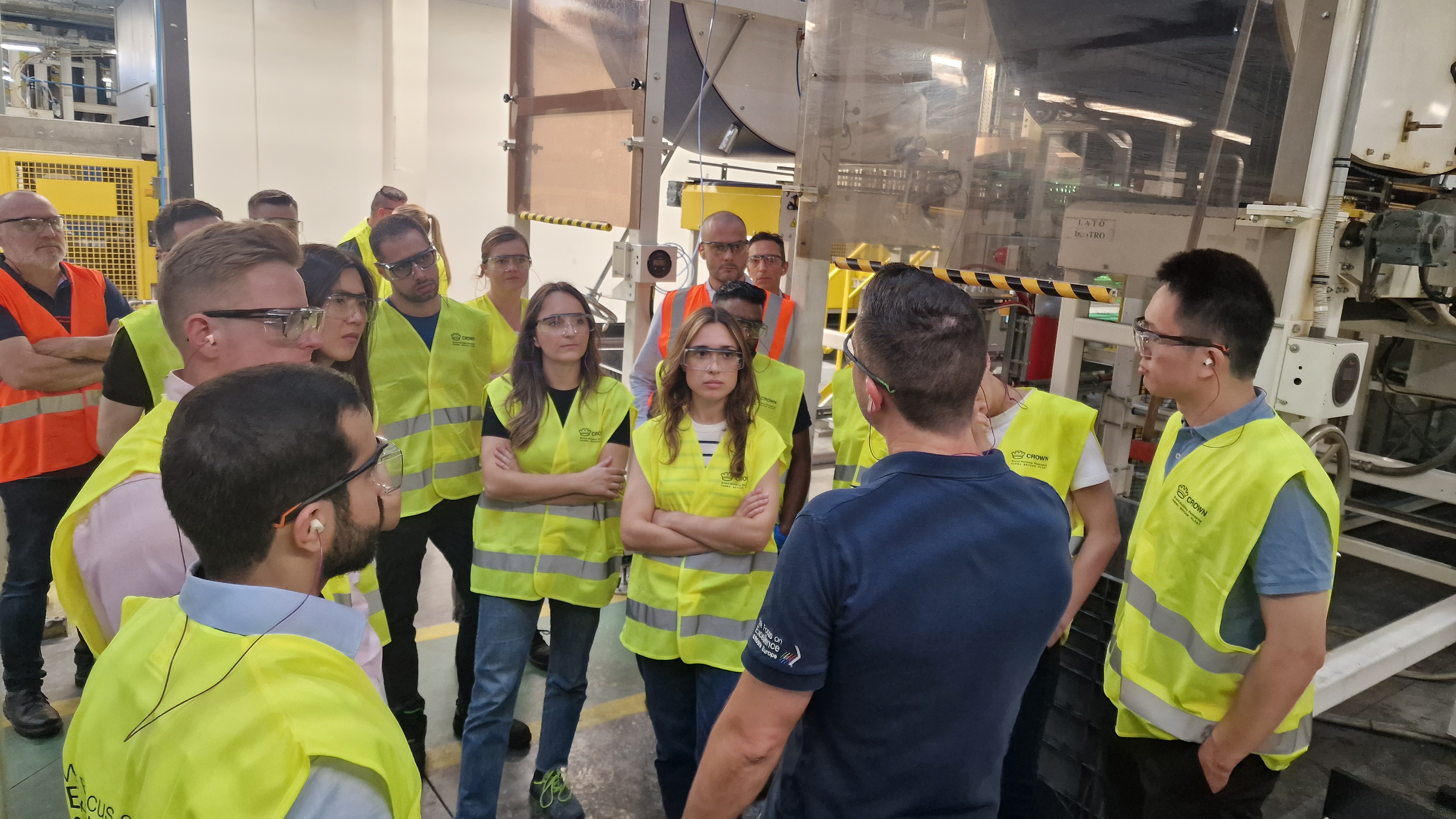 A group of people wearing high-visibility vests, safety glasses, and earplugs listening to a male-presenting person in a warehouse.