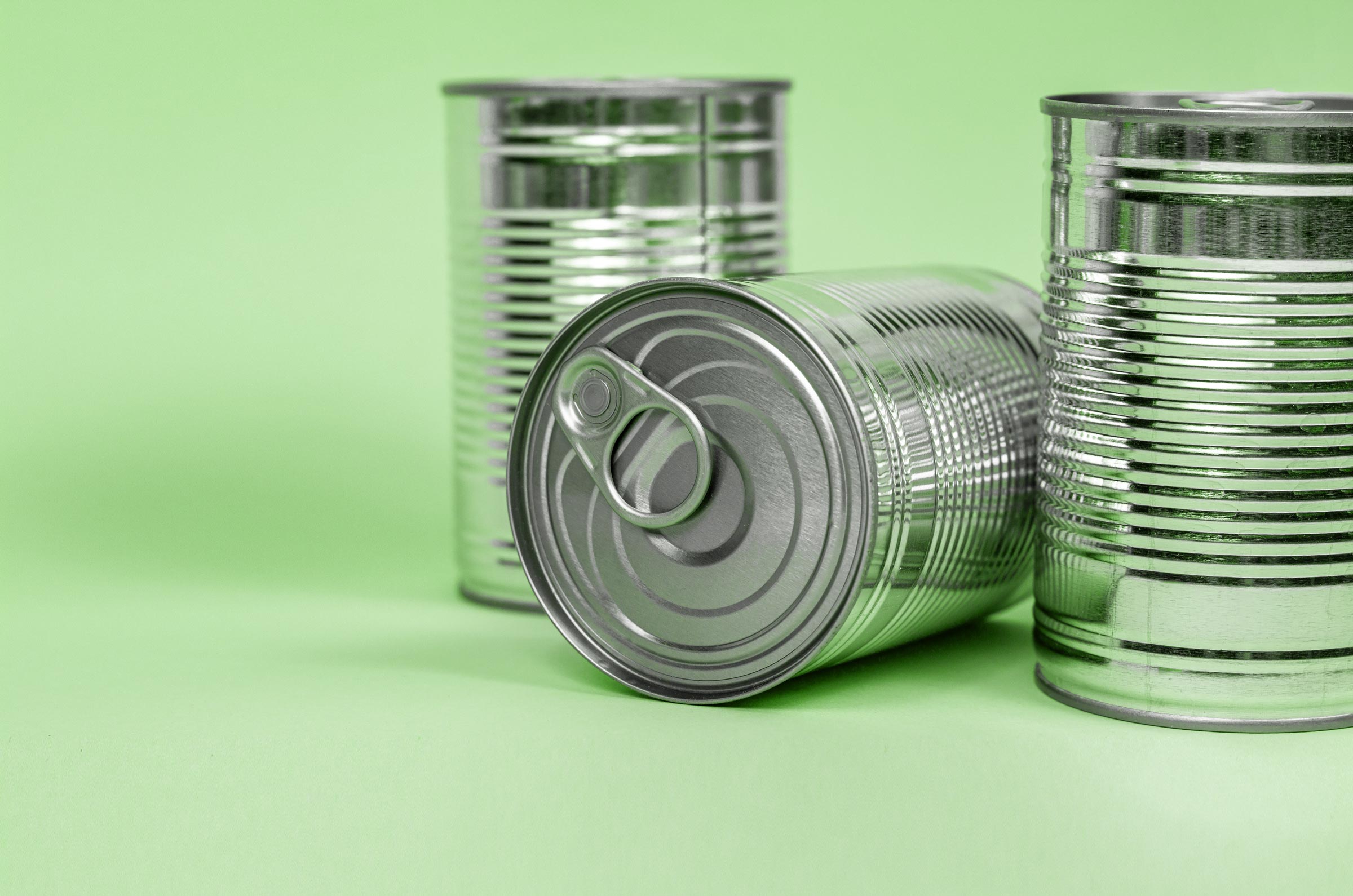 Three silver tin cans on a light green surface, one lying on its side showing a pull-tab lid.
