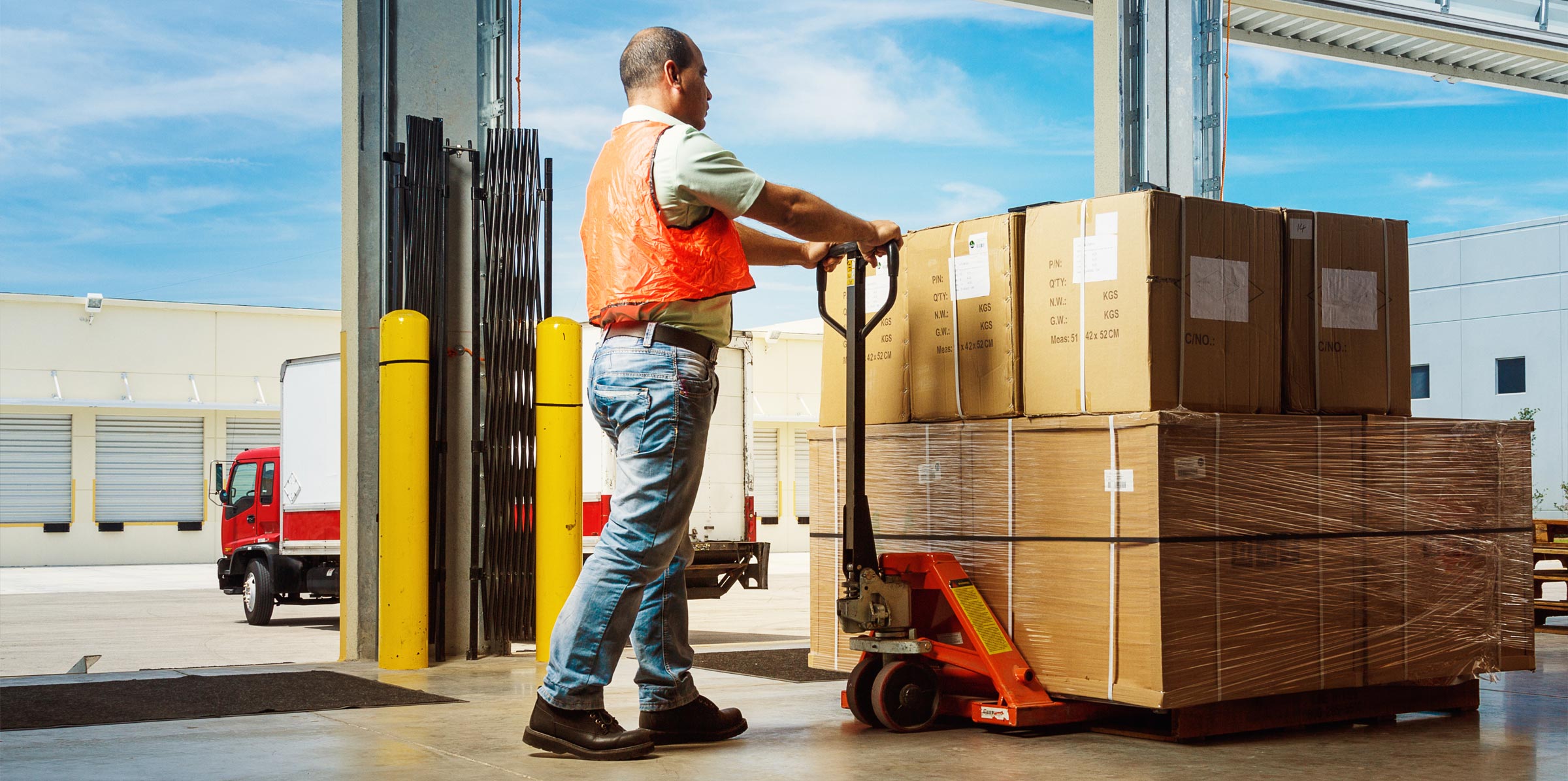 Worker using a pallet jack to move large stacked cardboard boxes in a warehouse loading area.
