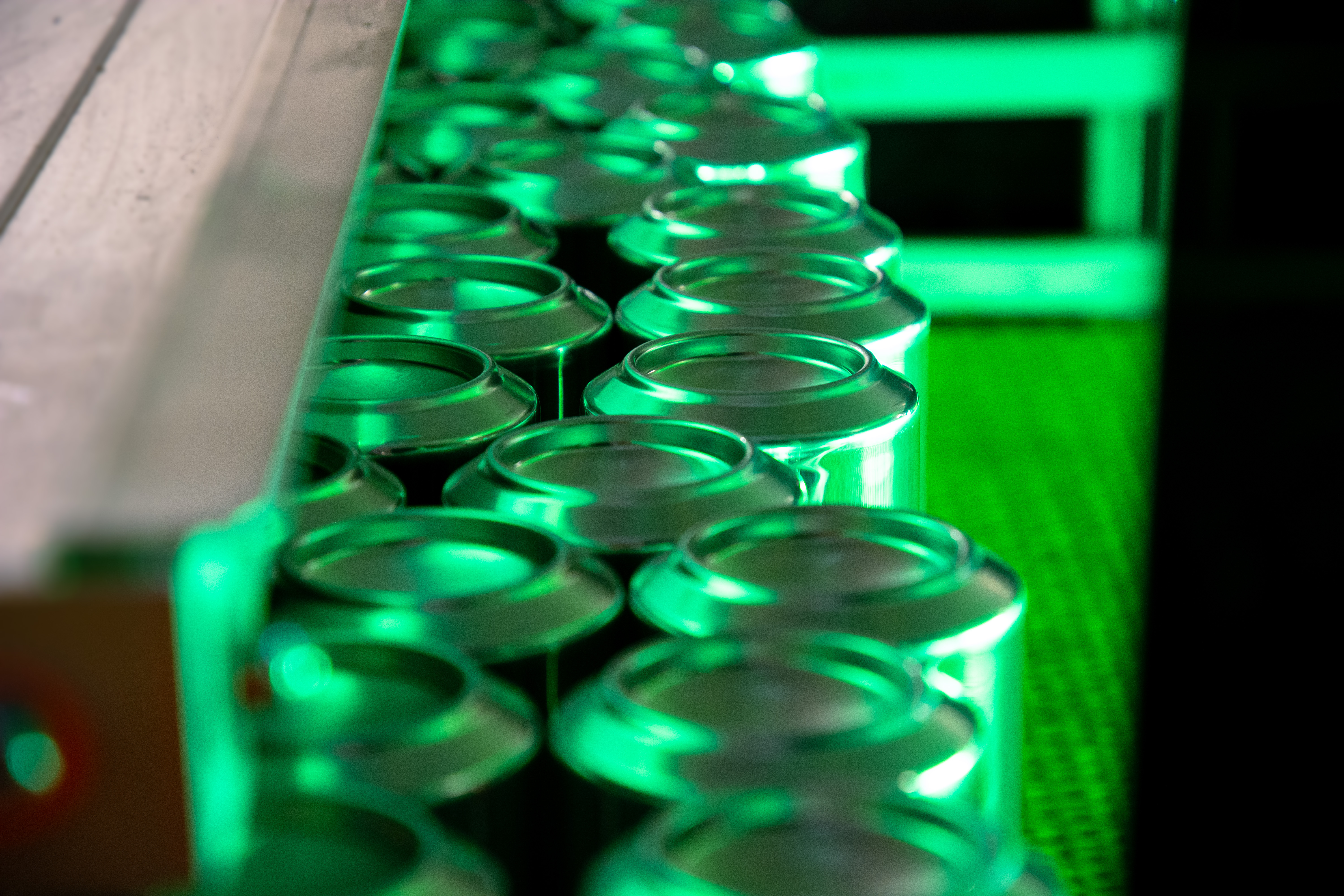 Close-up of empty aluminum beverage cans moving along a conveyor belt in a manufacturing facility, illuminated by green lighting.
