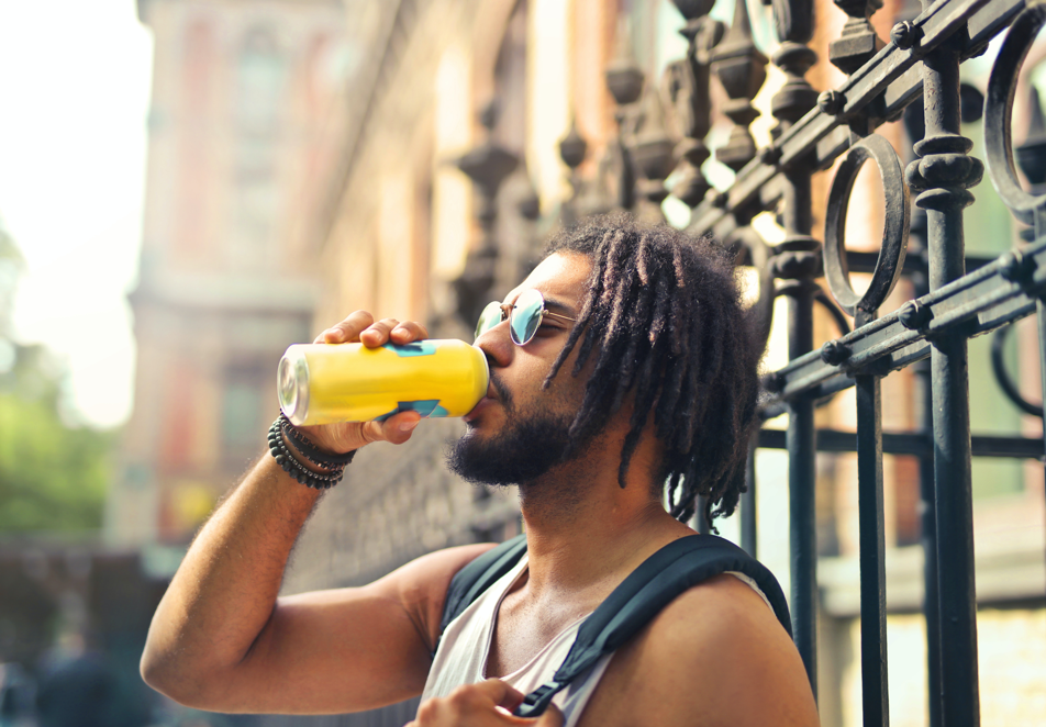 Wellness in a Can A medium-skinned man with short dreadlocks stands in front of a fence and drinks a can of juice.