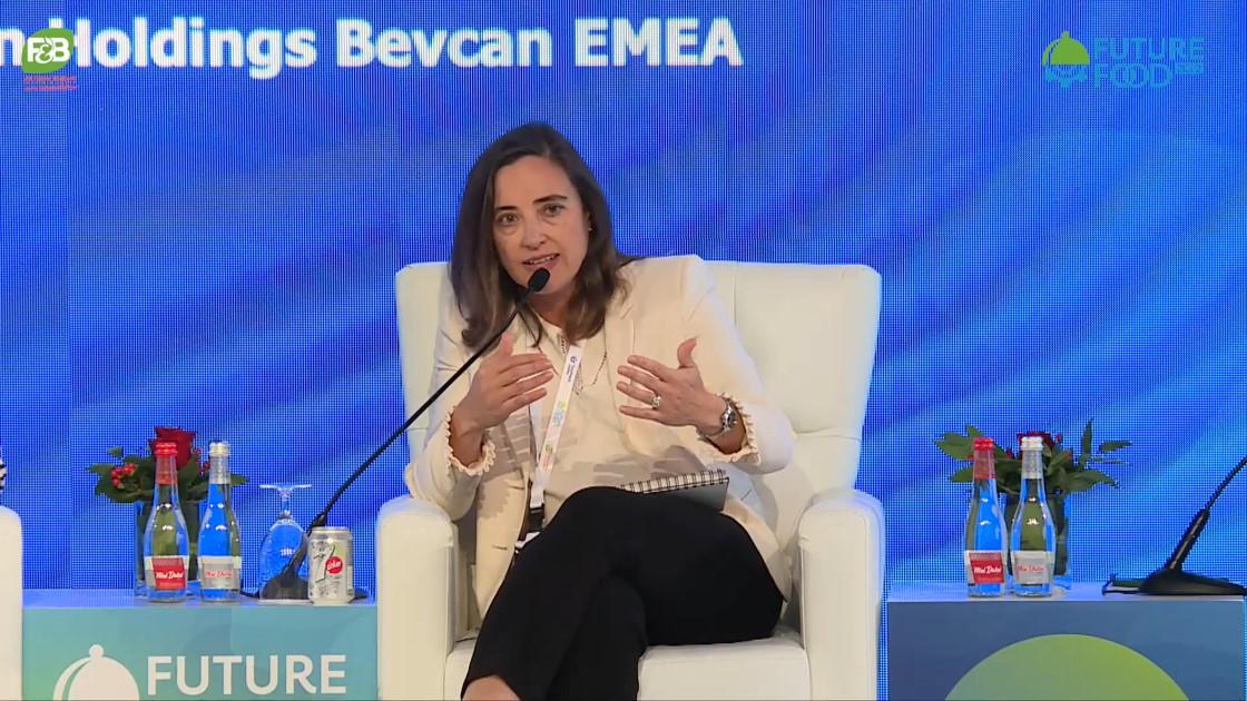A female-presenting person in business clothing sits in an office chair and speaks into a microphone on a stage.