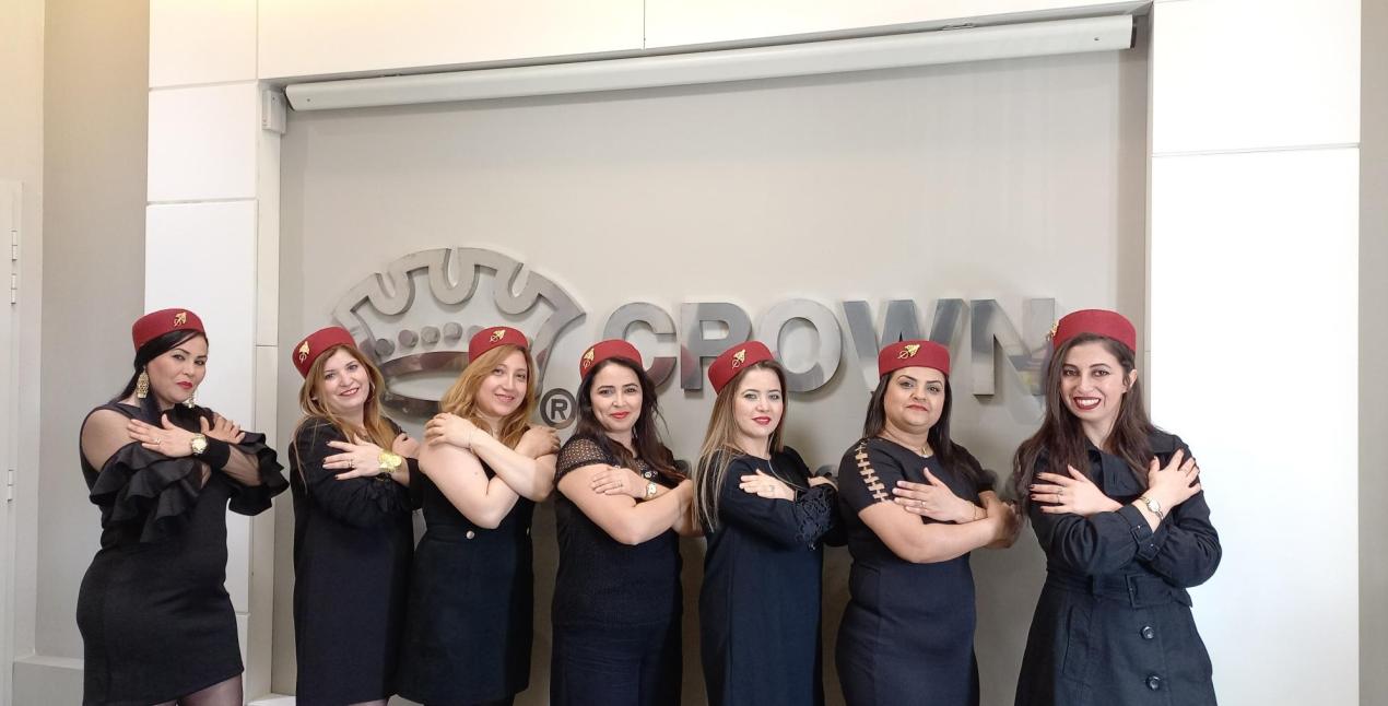 A group of women in navy dresses and red hats stand in front of the Crown Cork logo. They have their arms crossed in the symbol for 'equity'.
