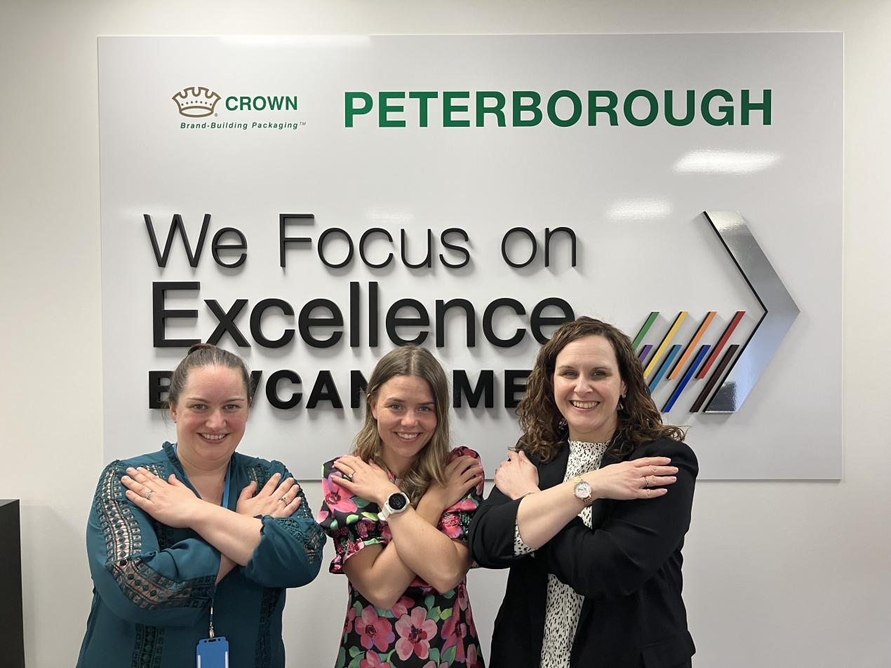 Three light-skinned women stand in front of a wall mural for Crown Peterborough. They have their arms crossed in the symbol for 'equity'.