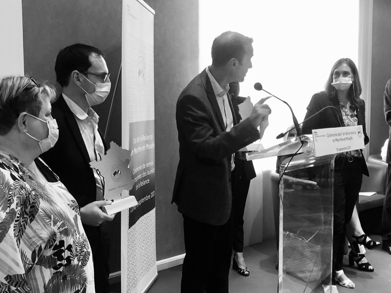 Five people stand around a podium where one man is speaking. Another man stands off to the side, holding an award.