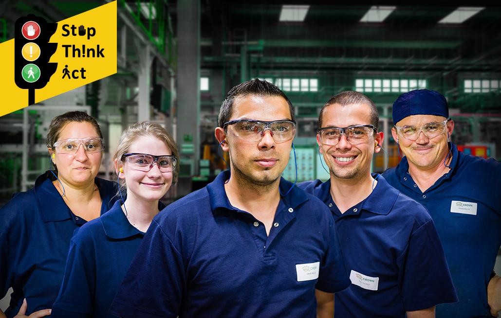 A group of Crown employees stand in a warehouse wearing safety glasses and earplugs with a sign that says "Stop, think, act."