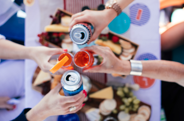 Four people off-screen clink a combination of glasses and aluminum cans together.