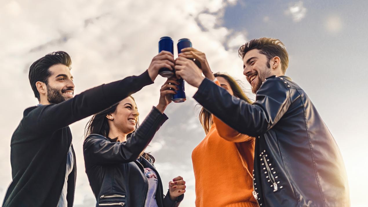 Two masc-presenting people and two fem-presenting people stand in a circle and 'cheers!' a set of blue cans.