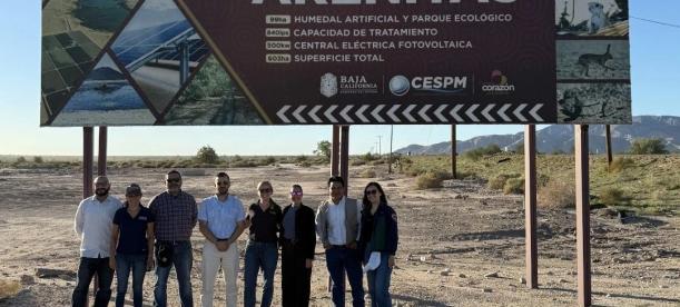 Group of people standing in front of a roadside sign for the Las Arenitas treatment plant, artificial wetland, and photovoltaic power facility in Baja California, Mexico.