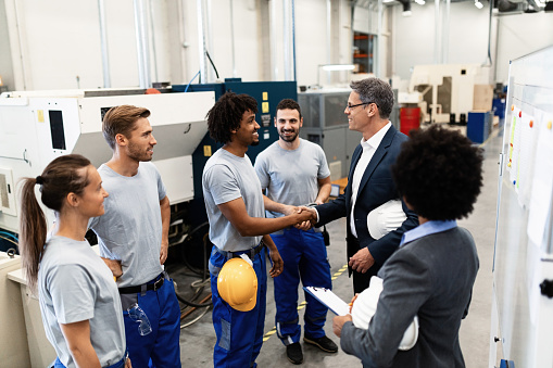 A male-presenting person in work clothing shakes hands with another male-presenting person in business clothing inside a warehouse.