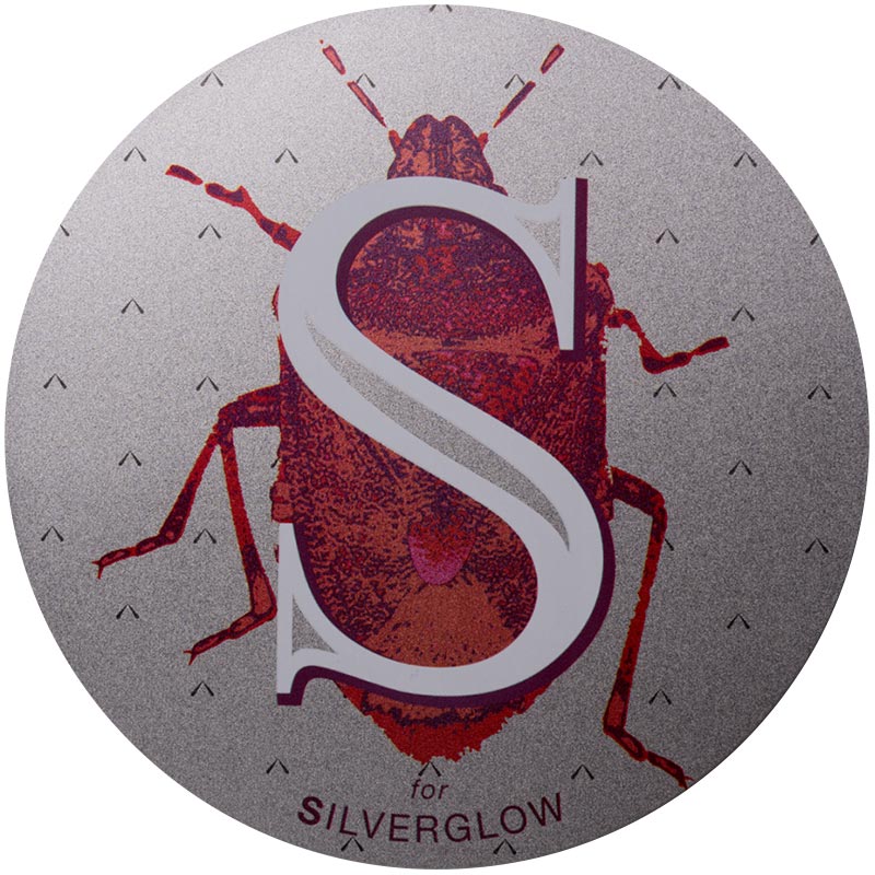 Silverglow-2x.jpeg A silver glow surface with a big letter "S" and a drawing of a red beetle behind the S.
