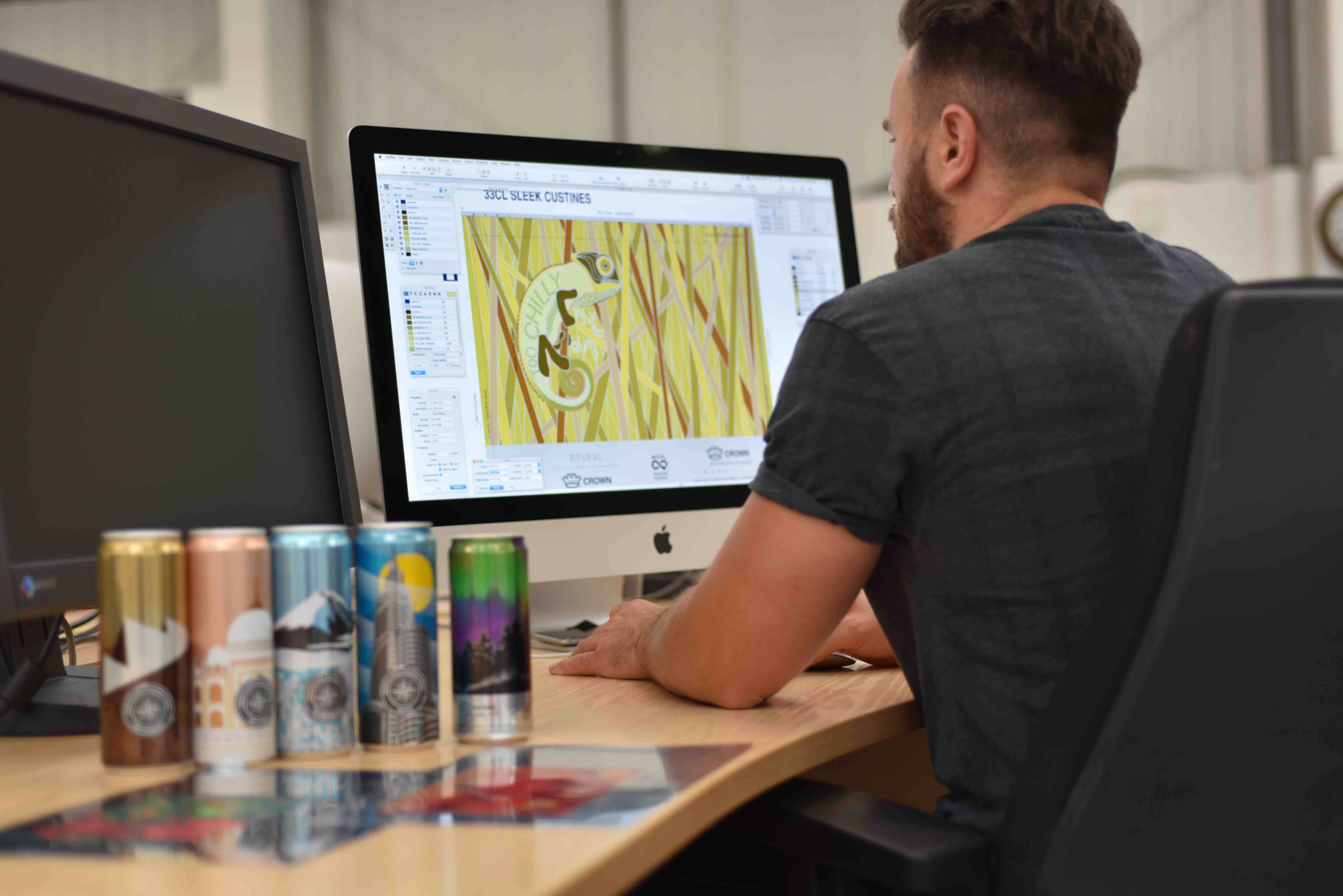 Male presenting person sits at a desk on a computer. Working on artwork for various cans. 