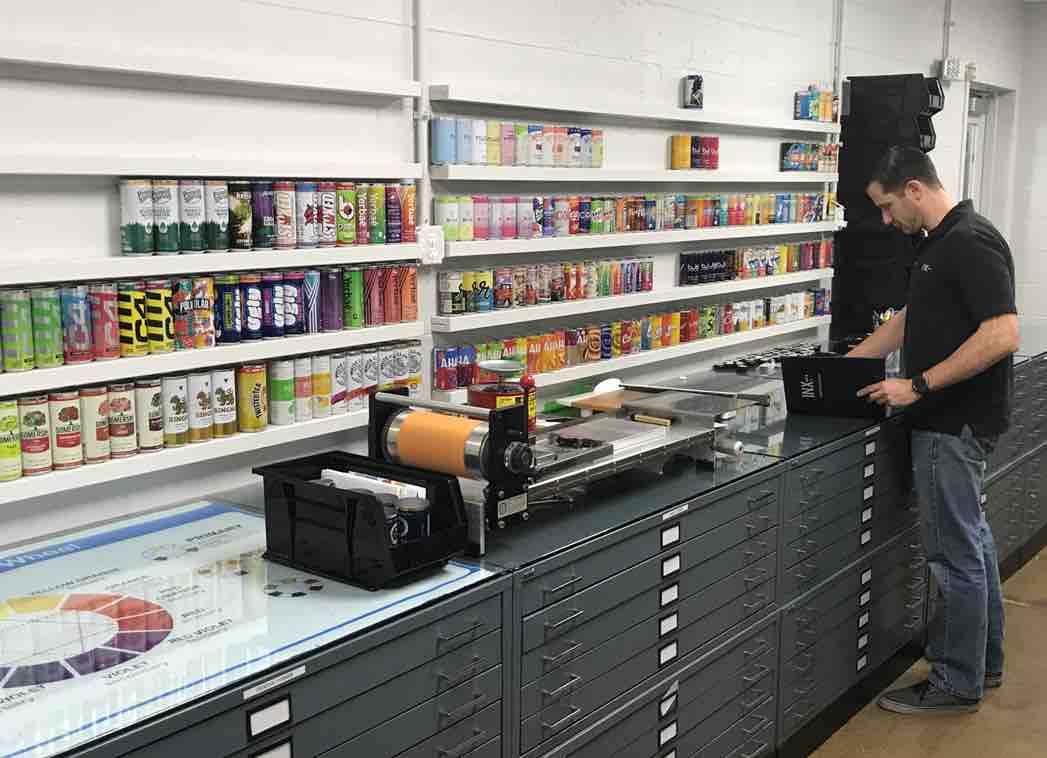 Male presenting person standing at a work station desk while matching ink samples. Shelves with various can displays sit on the wall. 