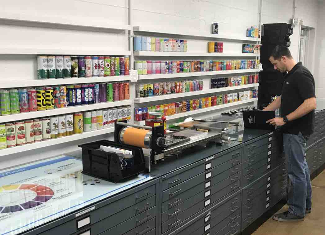 Male presenting person standing at a work station desk while matching ink samples. Shelves with various can displays sit on the wall.