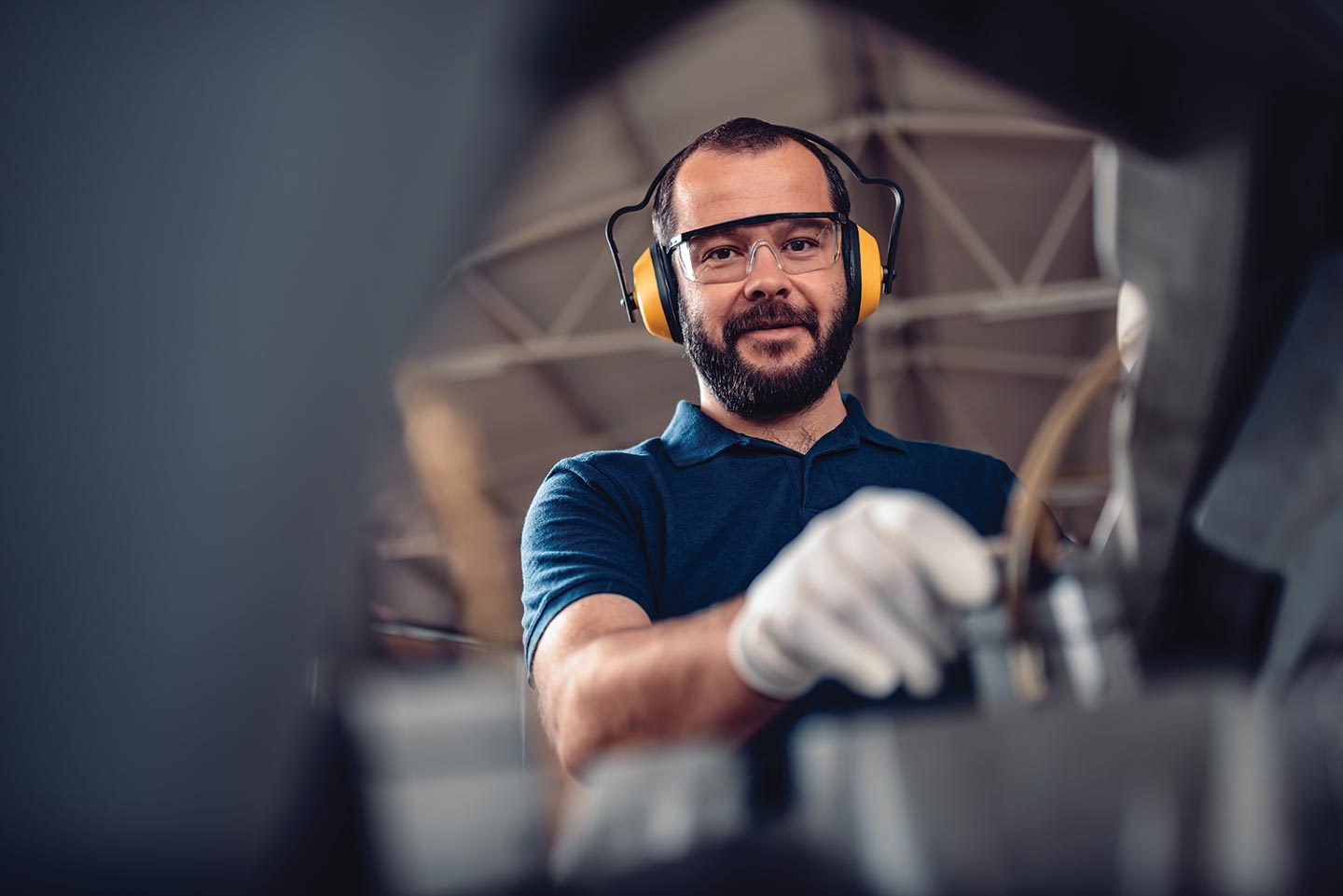 A male-presenting person wearing shop gloves, safety glasses, and ear protection smiles towards the camera while working in a shop.
