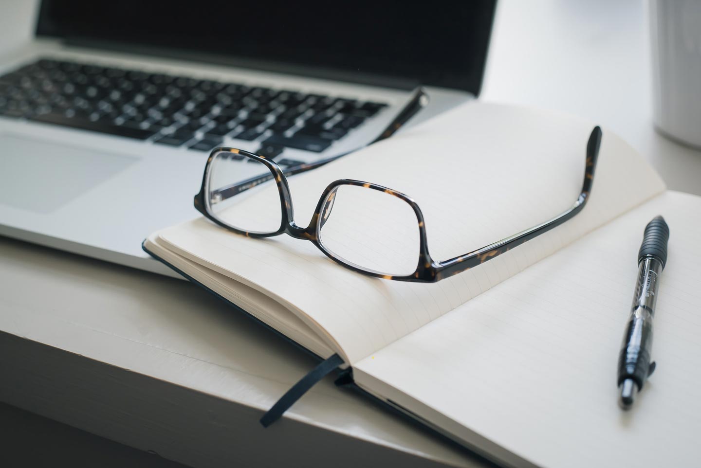 A pair of reading glasses sits on an open notebook next to a pen and a laptop on a desk.