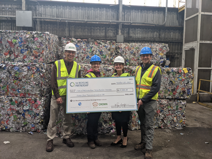 Milwaukee Area Recycling Facility Receives Check for Aluminum Beverage Can Capture Grant. Pictured (left to right) from the City of Milwaukee, Rick Meyers, sanitation services manager, and Samantha Longshore, resource recovery program manager, and from Waukesha County, Analiese Smith, recycling and solid waste supervisor, and Alan Barrows, land resources manager. Four people in hard hates and safety vests hold a large check while standing in front of large blocks of crushed cans.