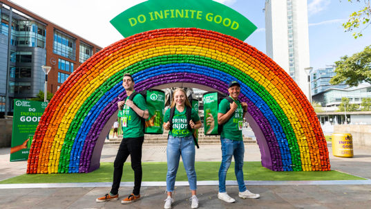 A group of people stand outdoors in front of a rainbow decoration made out of recycled aluminum cans.
