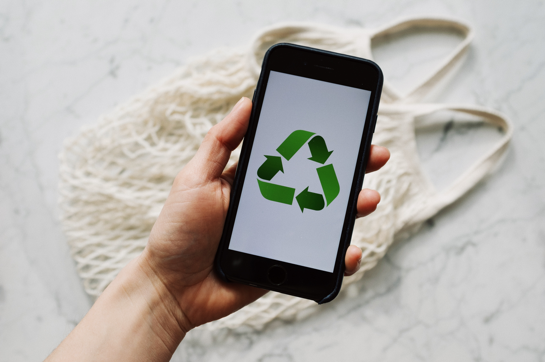 A person off-screen holds a smartphone with a green recycling icon.