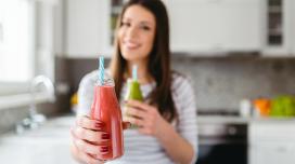 A female-presenting person appears to be handing a red smoothie in a clear glass towards the camera.