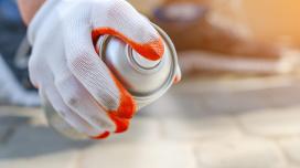 A person out-of-frame sprays an aerosol can downwards while outdoors and wearing protective gloves.