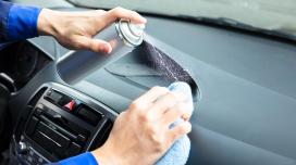 A person out of frame sprays an aerosol can onto the dashboard of a car while using a wipe to clean the dashboard.